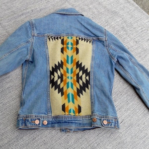May include: A blue denim jacket with a colorful geometric pattern on the back. The pattern is in shades of brown, yellow, blue, and black.