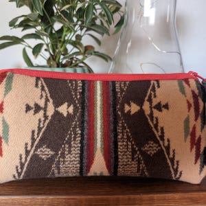May include: A rectangular, patterned pouch with a red zipper and trim. The pouch features a geometric design in shades of brown, beige, red, and green. A small leather loop is attached to the zipper. The pouch is likely made of a woven fabric.