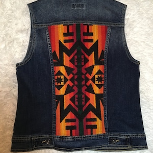 May include: A dark blue denim vest with a colorful geometric pattern on the back. The pattern is in shades of orange, red, black, and green.