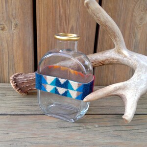 May include: A clear glass bottle with a gold top and a colorful patterned band around the middle. The bottle is sitting on a wooden surface next to a white antler.