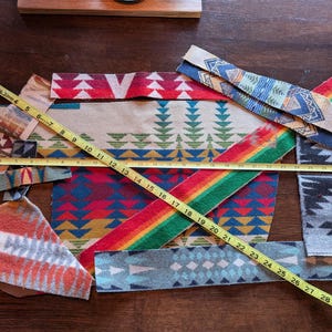 May include: Assorted fabric swatches with colorful geometric patterns, including red, blue, green, and orange. A yellow tape measure shows measurements up to 31 inches. The fabrics are arranged on a dark wooden surface.