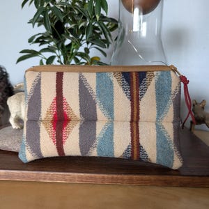 May include: A rectangular beige and patterned pouch with a zipper closure. The pattern includes blue, gray, red, and brown geometric shapes. The pouch is made of a woven fabric and has a brown zipper.