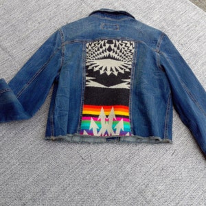 May include: A blue denim jacket with a colorful patterned fabric patch on the back. The patch features a rainbow design with geometric shapes and arrows.