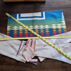 May include: A colorful textile with a geometric pattern in blue, green, red, and white, with a yellow measuring tape laid across it. The tape measures up to 26 inches. Several fabric scraps are also visible.