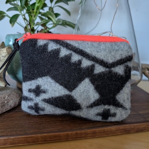 May include: A small, rectangular gray and black patterned zippered pouch. The pouch has a bright orange zipper and a black wrist strap. The design features geometric shapes and crosses.