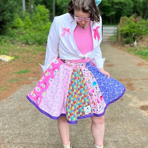 May include: A woman wearing a white button-down shirt with pink bows, a pink tank top, a colourful patchwork skirt, and beige ankle boots. She is wearing a blue bow in her hair and has on glasses.
