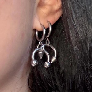May include: Two silver-toned circular hoop earrings are worn in the earlobe. Each hoop features a dangling horseshoe-shaped charm. One charm has two spherical ends, while the other charm has one spherical end and one pointed conical end.