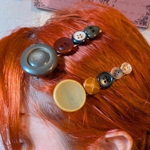 Op de afbeelding: Een persoon met felrood haar gestyled met verschillende haarclips met knopen. De clips bevatten knopen in verschillende maten en kleuren, waaronder bruin, zwart en crème. Grote oorringen en een zwarte top maken de look compleet.