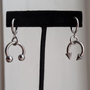 May include: A pair of silver-toned hoop earrings featuring dangling horseshoe-shaped charms. One charm is finished with two smooth spherical balls at the ends, while the other charm features two pointed conical spikes at the ends of the horseshoe.