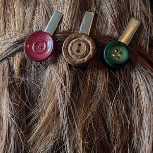 May include: Three hair clips with button-shaped accents. One is a deep red, another is brown, and the third is green. Each button is attached to a silver-colored metal clip, designed to hold hair in place. The clips are set against a backdrop of brown hair.