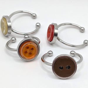 May include: Four button rings with silver-colored bands. The buttons are in various colors: yellow, red, orange, and brown. The rings are set against a white background.