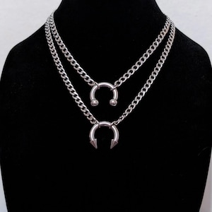 May include: Two silver-toned necklaces with curb chains. The top necklace features a horseshoe pendant with two small ball ends. The bottom necklace has a horseshoe pendant with two pointed cone ends. Both necklaces are displayed on a black mannequin.