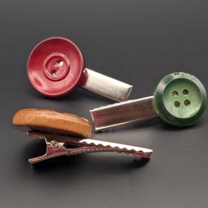 Può includere: Tre fermagli per capelli unici. Uno presenta un bottone rosso, un altro un bottone verde e il terzo un bottone in legno. Ogni bottone è attaccato a una clip in metallo argentato. Le clip sono disposte su uno sfondo scuro.