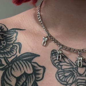 Könnte beinhalten: Eine silberne Halskette mit drei kleinen Zahnanhängern. Die Halskette wird am Hals einer Person getragen, wobei Tätowierungen von Blumen und einem Schmetterling auf der Haut sichtbar sind.
