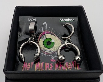 May include: Two silver-toned hoop earrings with dangling horseshoe-shaped charms in a black box. One charm has pointed spikes, the other has round balls. The display card features a green eye illustration, the text Luxe, Standard, and HOT NICHE NEUROTIC.