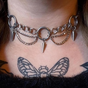 May include: A silver choker necklace with a punk rock aesthetic. The necklace features a series of small chains, large rings, and dangling cone-shaped spikes. The necklace is worn around the neck.