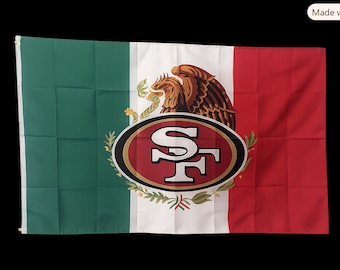 Puede incluir: Una bandera rectangular con los colores verticales verde, blanco y rojo de la bandera mexicana. En el centro de la franja blanca hay un águila marrón sobre un cactus, superpuesta con el logotipo rojo y dorado de los San Francisco 49ers con las letras SF.