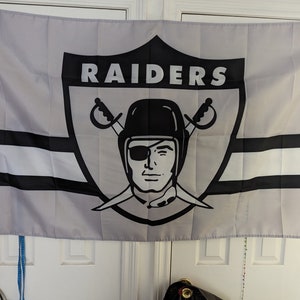 May include: A gray and black flag with the word "RAIDERS" in white letters on a black shield. The shield features a black and white illustration of a pirate with an eyepatch and crossed swords.