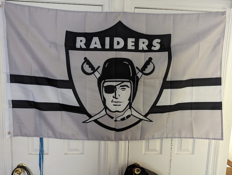 May include: A gray and black flag with the word "RAIDERS" in white letters on a black shield. The shield features a black and white illustration of a pirate with an eyepatch and crossed swords.