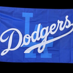 May include: A rectangular blue flag featuring the white script text Dodgers overlaid on a large, light blue block letter LA logo. The flag has visible horizontal and vertical fold lines across the fabric surface and metal grommets on the left side.