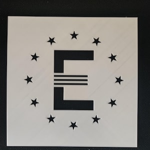 Może przedstawiać: Biały kwadrat z czarną literą "E" pośrodku, otoczony okręgiem czarnych gwiazd. Litera "E" ma trzy poziome linie przez środek.