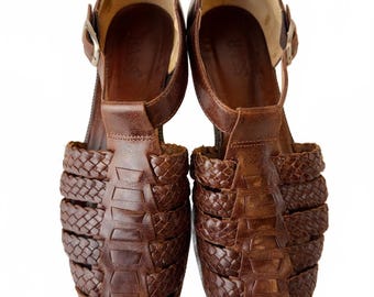 May include: A pair of brown leather huarache-style sandals featuring a woven lattice design across the vamp, a closed rounded toe, and an adjustable ankle strap with a silver-tone metal buckle. The interior footbed is stamped with the Bass brand name.
