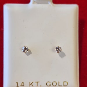 May include: A pair of small, round, clear diamond earrings set in 14 karat gold. The earrings are displayed on a white card with the words "14 KT. GOLD" printed on it.