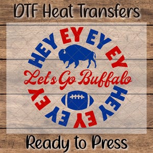 May include: DTF Heat Transfers design with the text "HEY EY EY" in blue and red, surrounding a blue buffalo and a football. The words "Let's Go Buffalo" are in red script. The text "Ready to Press" is at the bottom.