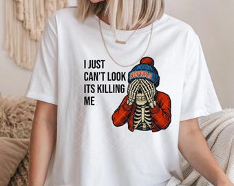 May include: White t-shirt with the text "I just can't look its killing me." The graphic shows a skeleton wearing a blue and red Buffalo Bills beanie and a red plaid jacket, covering its eyes with its hands.
