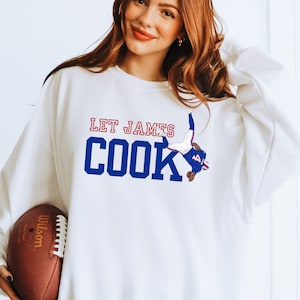 May include: White sweatshirt with a blue and red graphic of a football player in a diving pose. The text "LET JAMES COOK" is printed in blue on the sweatshirt.
