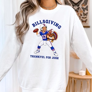 May include: A white long-sleeve sweatshirt featuring a graphic of a football player wearing a blue jersey with the number 17 and a helmet, holding a football and a turkey. The text reads BILLSGIVING and THANKFUL FOR JOSH.