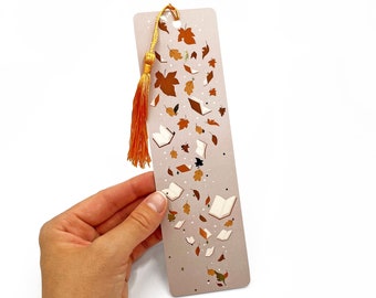 May include: A light brown bookmark with a gold tassel. The bookmark features a design of open books and autumn leaves falling.