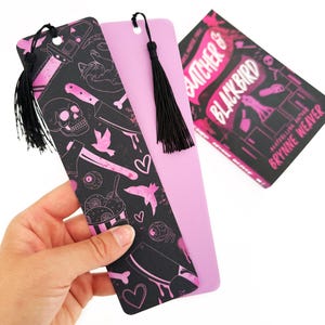 May include: A black and pink bookmark with a pink tassel. The bookmark features a skull, knives, hearts, and other spooky imagery. The bookmark is paired with a pink book with the title "Slaughter of Blackbird" on the cover.