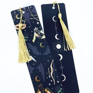 May include: Two black bookmarks with gold accents and tassels. One bookmark features a crescent moon, stars, mountains, and a bow and arrow. The other bookmark features a crescent moon, stars, and a sun.