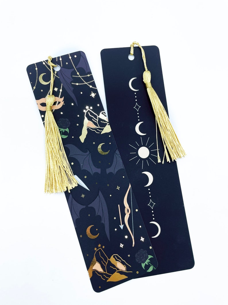 May include: Two black bookmarks with gold accents and tassels. One bookmark features a crescent moon, stars, mountains, and a bow and arrow. The other bookmark features a crescent moon, stars, and a sun.