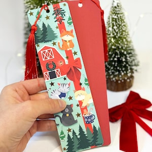 May include: A red and green Christmas-themed bookmark with a red tassel. The bookmark features a red barn, a green Christmas tree, and a white cat with a red bow. The bookmark is on a white background.