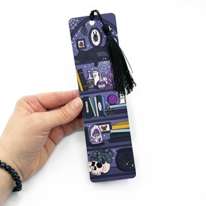 May include: Purple bookmark with a black tassel featuring a bookshelf with various magical items, including books, potions, crystals, a skull, a cat, and a spider.