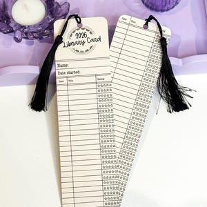 May include: Two library card bookmarks with black tassels. The bookmarks have a vintage design with lines for writing and the text "2026 Library Card". The background includes purple decorative items.