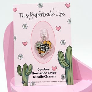 Puede incluir: Un charm de acrílico transparente con un sombrero de vaquero y las palabras "Cowboy Romance Lover" en una tarjeta rosa. La tarjeta tiene un diseño floral y de corazón con el texto "This Paperback Life".