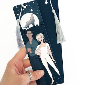 Może przedstawiać: Para czarnych zakładek z wilkiem wyjącym do księżyca, gwiazdami i parą stojącą przed lasem. Zakładka ma biały frędzl.