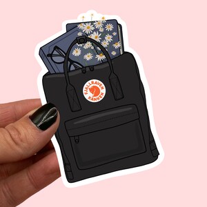 Könnte beinhalten: Schwarzer Fjällräven Kånken Rucksack mit einem weißen Reißverschluss und einem weißen Fjällräven Logo. Der Rucksack ist mit Büchern und weißen Gänseblümchen gefüllt.