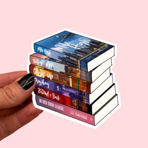 Può includere: Una pila di sei libri con pagine bianche e copertine colorate. I titoli includono "Mile High", "The Right Move" e "In Her Own League". Il nome dell'autore, Liz Tomforde, è visibile sulle copertine. I libri sono tenuti su uno sfondo rosa.