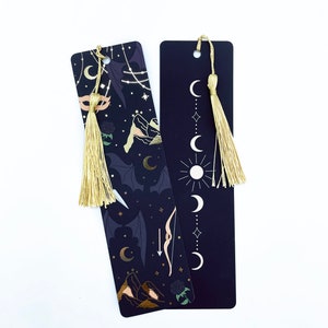 May include: Two black bookmarks with gold accents and tassels. One bookmark features a moon and star design, while the other has a more intricate design with mountains, a bow and arrow, and a crescent moon.