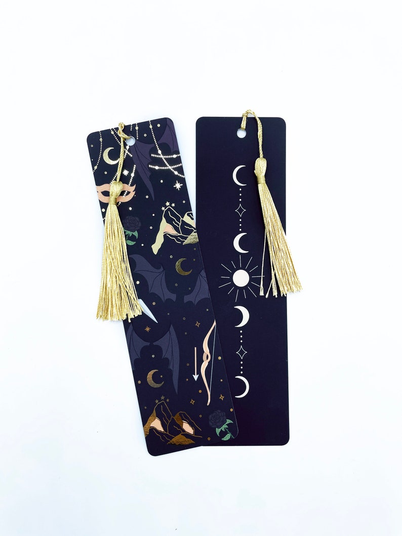 May include: Two black bookmarks with gold accents and tassels. One bookmark features a moon and star design, while the other has a more intricate design with mountains, a bow and arrow, and a crescent moon.