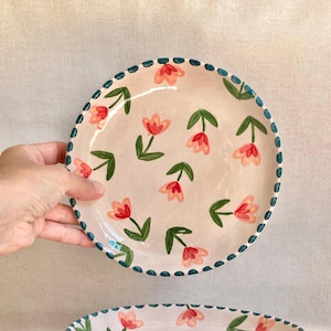 Peut inclure: Deux assiettes en céramique avec un motif floral. Les assiettes ont un fond rose clair avec des fleurs rouges et roses et des feuilles vertes. Les bords sont décorés d'un motif pointillé bleu foncé.