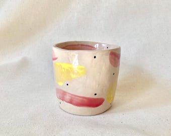 May include: A hand-built ceramic cup with a light beige base, featuring abstract brushstrokes in shades of pink and yellow, accented by small, scattered dark blue dots. The cup has an irregular, organic shape with a smooth, glossy finish.