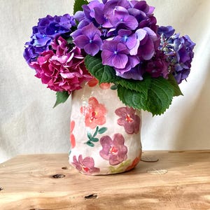 May include: A cylindrical ceramic vase with a light pink base featuring hand-painted red, orange, and purple floral motifs. The vase is filled with a large, dense bouquet of vibrant purple, blue, and magenta hydrangea flowers with green leaves.