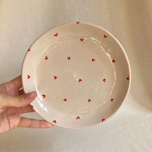 Pode incluir: Um prato de cerâmica feito à mão com uma base rosa claro e pequenos desenhos de corações vermelhos espalhados pela superfície. O prato tem uma borda ligeiramente irregular, dando-lhe um toque rústico e artesanal. Perfeito para servir comida ou como peça decorativa.