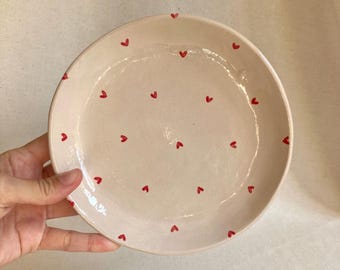 May include: A handmade ceramic plate with a pale pink base and small red heart designs scattered across the surface. The plate has a slightly irregular edge, giving it a rustic, artisanal feel. Ideal for serving food or as a decorative piece.