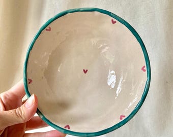 May include: A small, off-white ceramic bowl with a teal rim and small pink hearts painted around the edge. A single pink heart is in the centre of the bowl. The bowl is held by a hand.